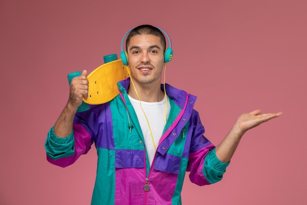 Free photo front view young male in colorful coat listening to music holding skateboard on pink desk