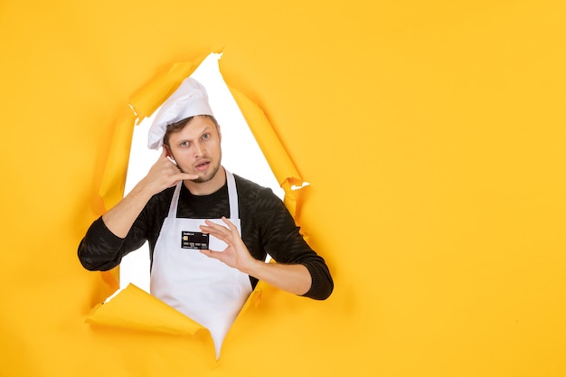 Front view young male cook in white cape holding black bank card on the yellow background model white color cuisine job man food money