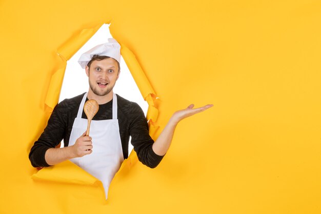 Front view young male cook in white cape holding wooden spoon on a yellow background color kitchen photo cuisine job white man food