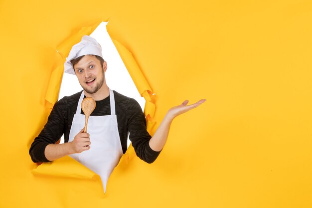 Front view young male cook in white cape holding wooden spoon on the yellow background color kitchen photo cuisine job white man food