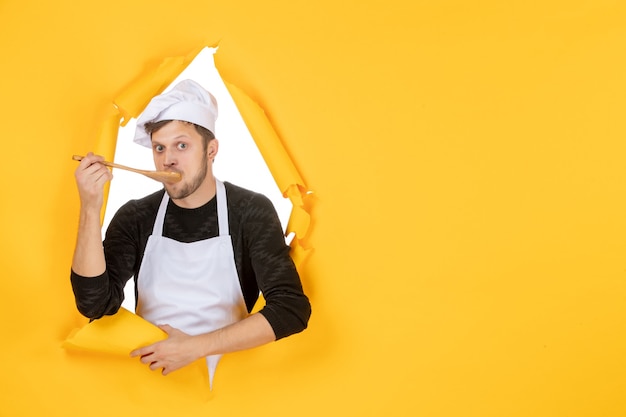 Free photo front view young male cook in white cape tasting wooden spoon on yellow background kitchen photo food cuisine job white color man