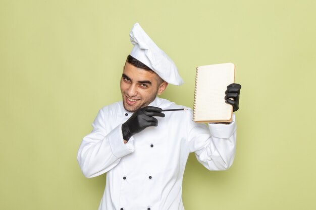 Front view young male cook in white cook suit holding notepad and smiling on green