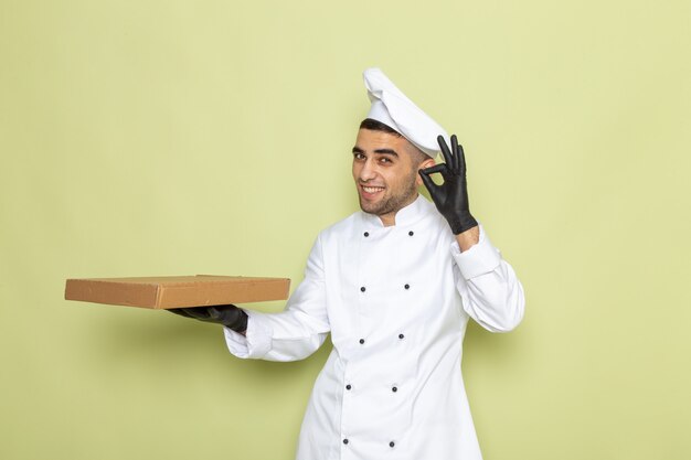 Front view young male cook in white cook suit wearing black leather gloves holding box on green