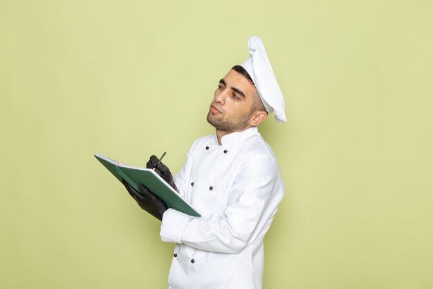 Front view young male cook in white cook suit wearing dark gloves and writing down notes on green