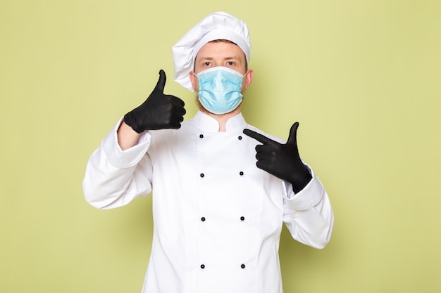 Free photo a front view young male cook in white cook suit white head cap in black gloves blue protective mask showing awesome sign