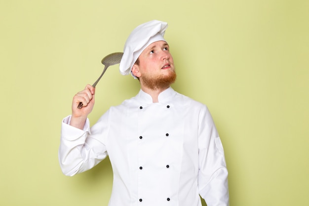 Free photo a front view young male cook in white cook suit white head cap holding big silver spoon thinking