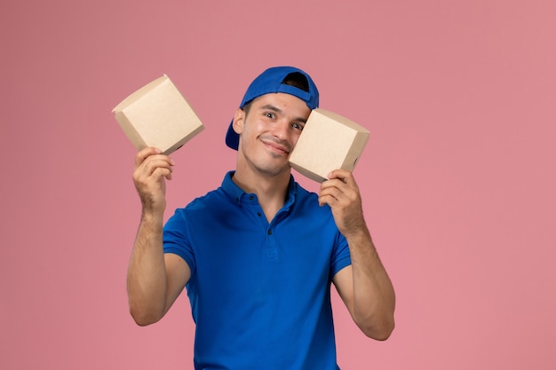 Front view young male courier in blue uniform cape holding little delivery food packages on light pink wall