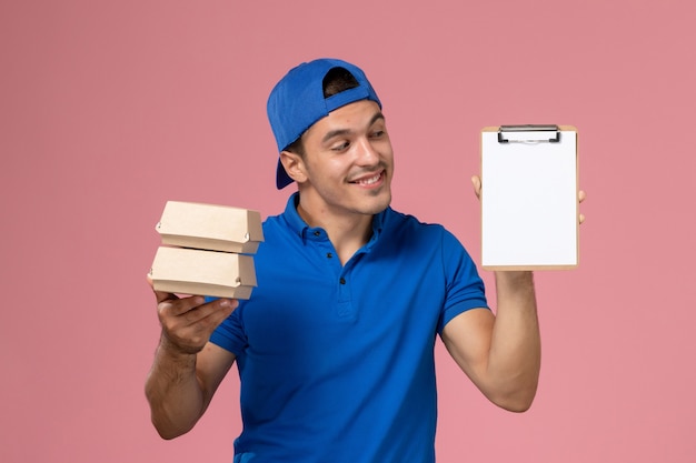 Free photo front view young male courier in blue uniform cape holding little delivery food packages with notepad and smiling on light pink wall