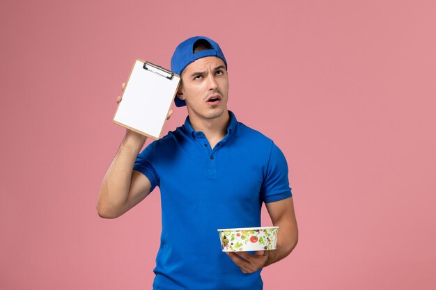 Front view young male courier in blue uniform cape holding notepad and round delivery bowl just thinking on pink wall