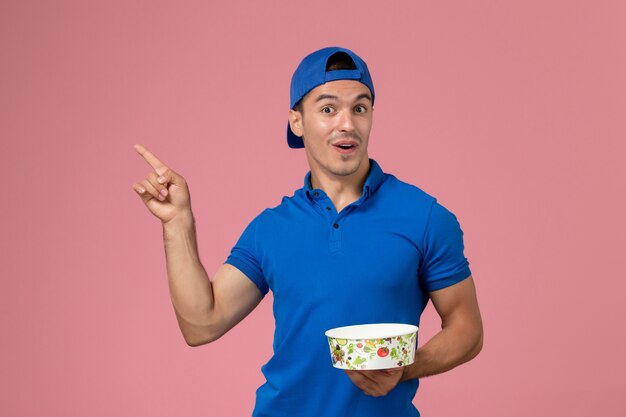 Front view young male courier in blue uniform cape holding round delivery bowl on light pink wall