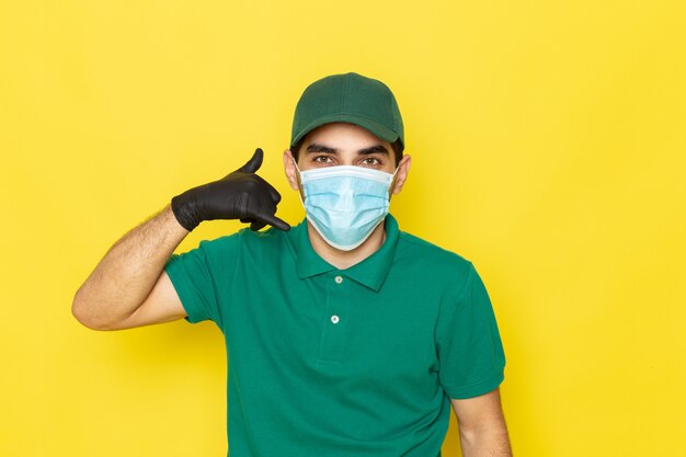 Front view young male courier in green shirt green cap black gloves showing phone call sign on yellow