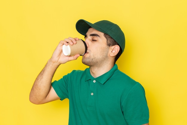 Front view young male courier in green shirt green cap drinking coffee on yellow