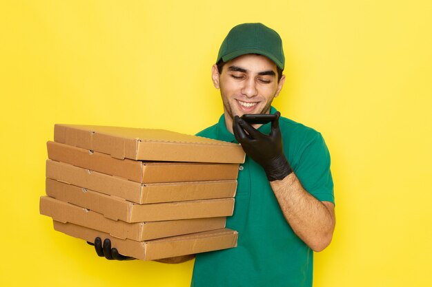 Front view young male courier in green shirt green cap holding delivery boxes and talking on phone smiling on yellow