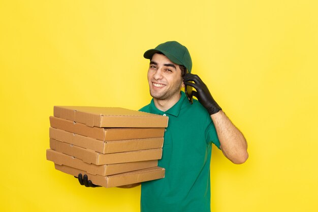 Front view young male courier in green shirt green cap holding delivery boxes and talking on phone on yellow