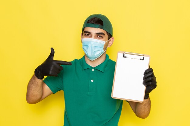 Front view young male courier in green shirt green cap holding notepad on yellow