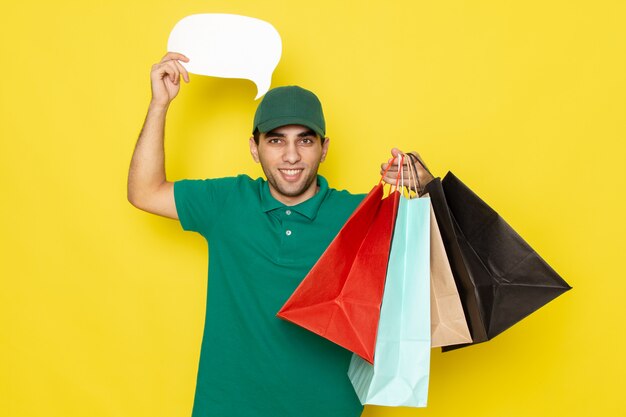 Front view young male courier in green shirt green cap holding shopping packages and white sign