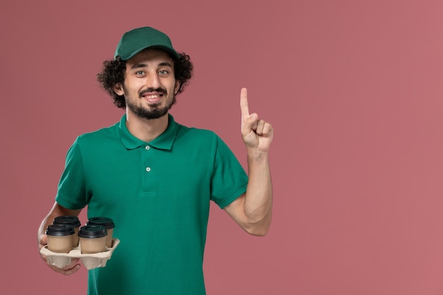 Free photo front view young male courier in green uniform and cape holding brown delivery coffee cups on the pink desk service uniform delivery worker