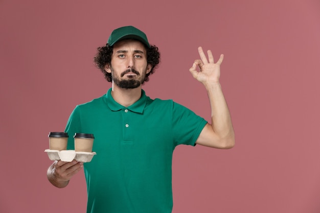 Front view young male courier in green uniform and cape holding delivery coffee cups on pink background service job uniform delivery