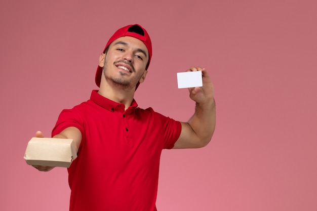 Front view young male courier in red uniform cape holding little delivery package with white card on pink background.