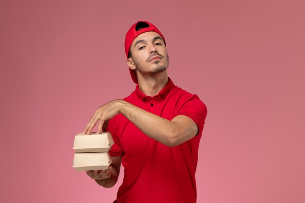 Front view young male courier in red uniform cape holding little packages with food on light-pink background.