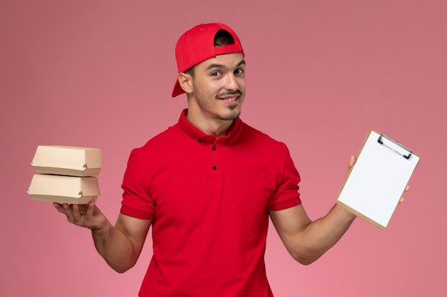 Front view young male courier in red uniform cape holding little packages with food and notepad on light-pink background.