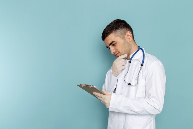 Front view of young male doctor in white suit with blue stethoscope holding notepad with thinking expression