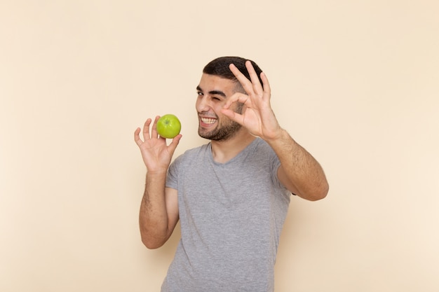 Front view young male in grey t-shirt and blue jeans smiling and holding green apple on beige