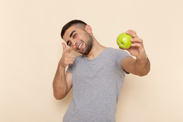 Front view young male in grey t-shirt and blue jeans smiling and holding green apple on beige