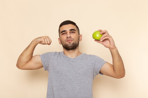 Front view young male in grey t-shirt holding apple and flexing on beige