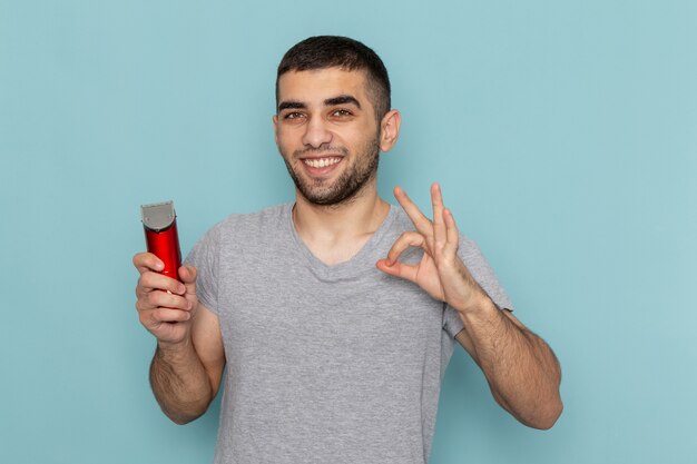 Front view young male in grey t-shirt holding electric razor showing alright sign on ice-blue beard foam