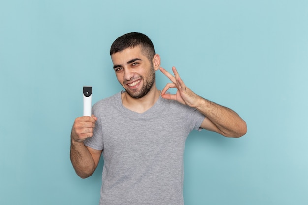 Front view young male in grey t-shirt holding electric razor with smile on the blue