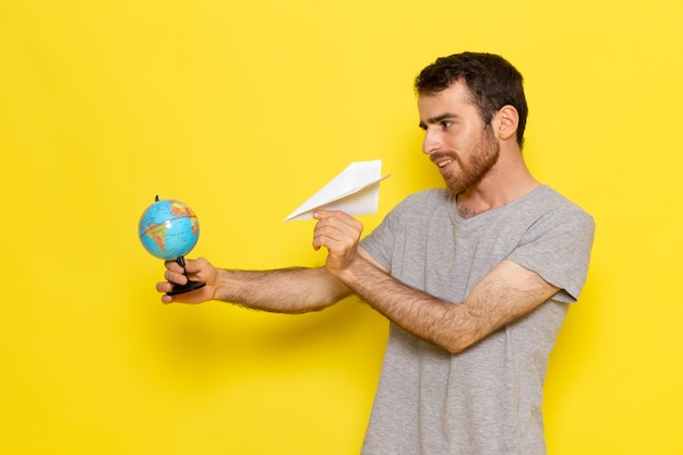 Free Photo a front view young male in grey t-shirt holding little globe and paper plane on the yellow wall man color model