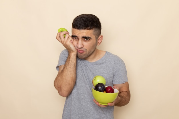 Front view young male in grey t-shirt holding plate with fruits biting apple having severe toothache on beige