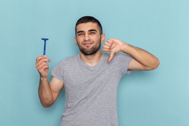 Front view young male in grey t-shirt holding razor and showing unlike sign on the blue