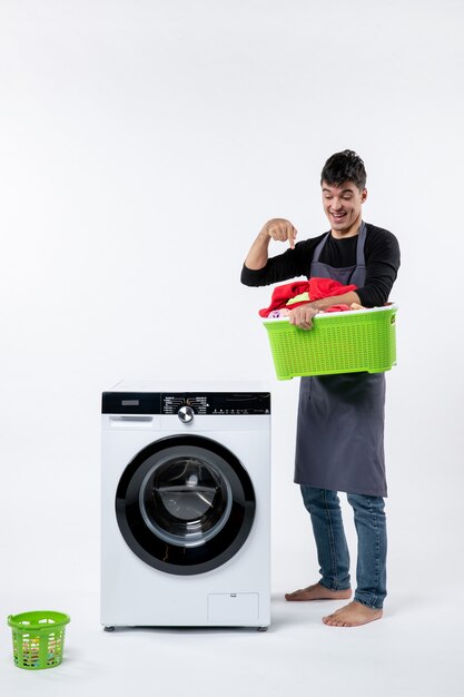 Front view of young male holding green basket with dirty clothes on the white wall