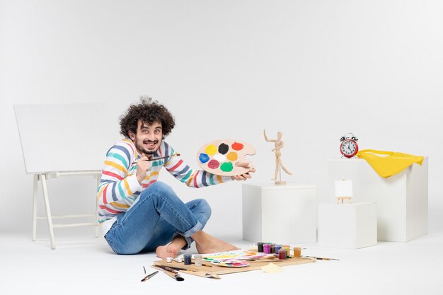 Front view of young male holding paints and tassel for drawing on white wall