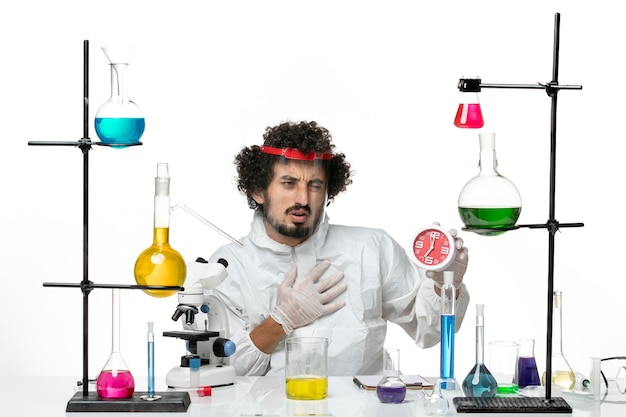 Front view young male scientist in special suit with protective helmet holding clock on light-white wall