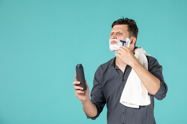 front view young male shaving his face with razor on blue background