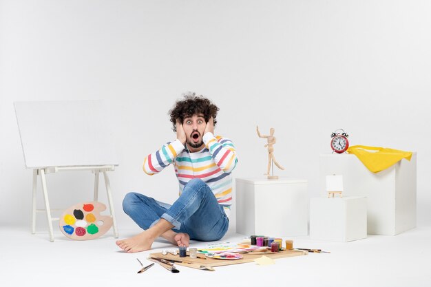 Front view of young male sitting around paints and drawings on the white wall