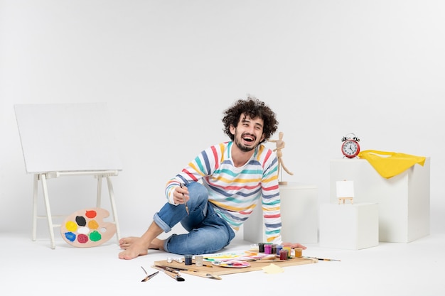 Free photo front view of young male sitting around paints and drawings on white wall