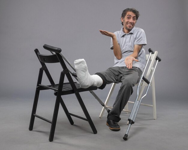 Front view young male sitting with broken foot and crutches on grey floor accident twist broken foot leg pain