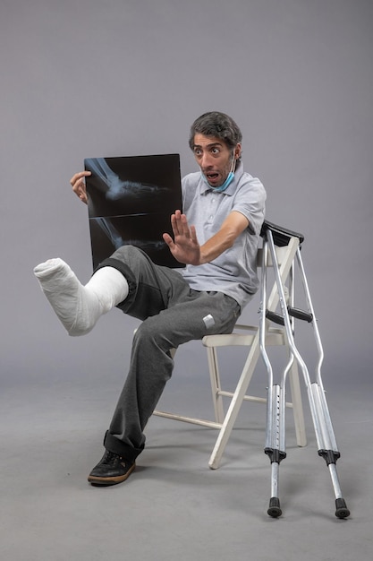 Front view young male sitting with broken foot and holding x-ray on the grey wall pain leg accident foot twist male