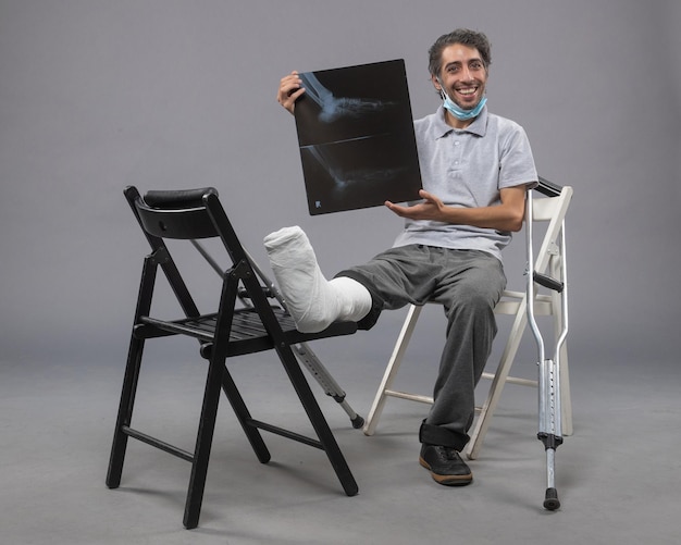 Front view young male sitting with broken foot and holding x-ray of it on grey floor twist pain foot leg broken male accident