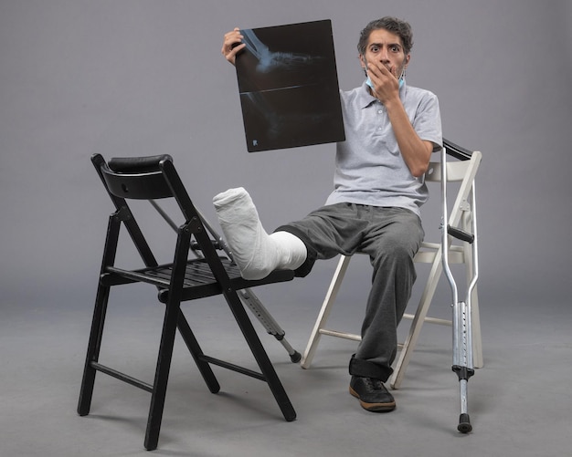 Front view young male sitting with broken foot and holding x-ray of it on grey wall medicine twist male pain foot legs broken