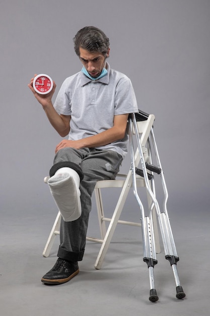 Free photo front view young male sitting with broken foot tied bandage and holding clock on grey desk twist pain foot accident leg male