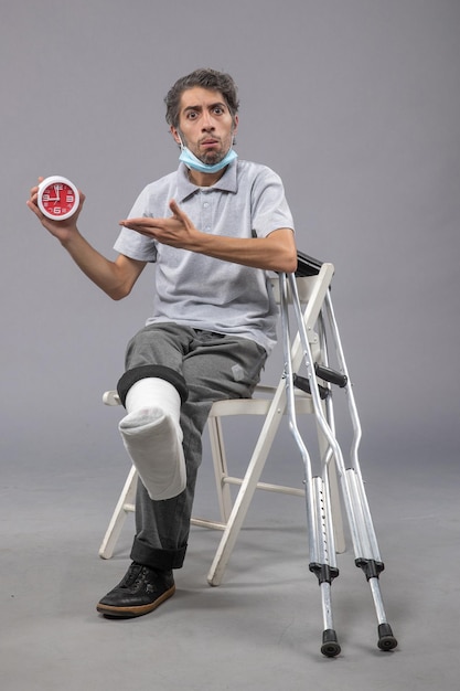 Front view young male sitting with broken foot tied bandage and holding clock on the grey wall foot twist accident pain leg male