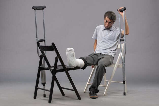 Front view young male sitting with broken foot and using crutches on a grey wall foot twist broken pain accident legs