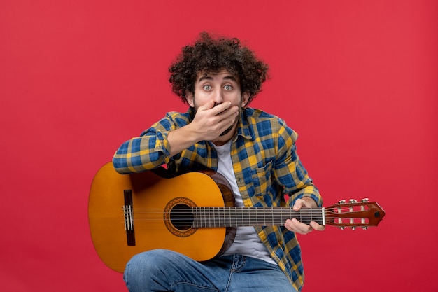 Front view young male sitting with guitar on a red wall play music performance musician color applause live concert