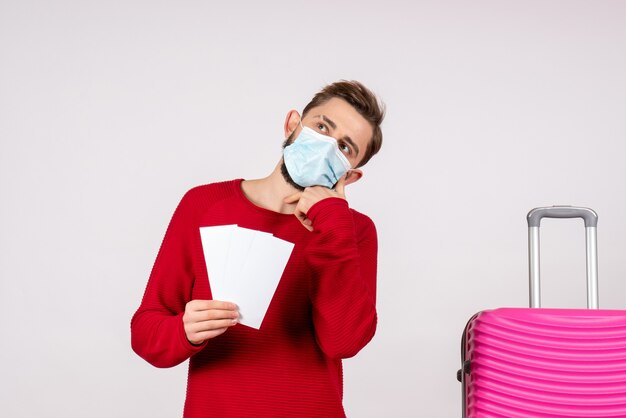 Front view young male in sterile mask holding tickets on white wall voyage covid- trip vacation emotion flight color