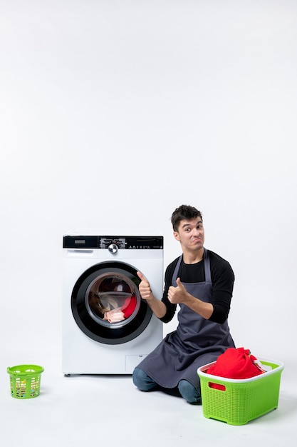 Front view of young male washing clothes with the help of washing machine on the white wall